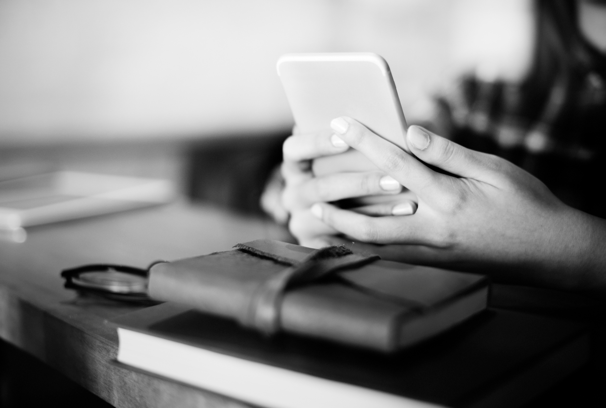 Person holding a smartphone at a desk with a closed leather notebook and glasses nearby