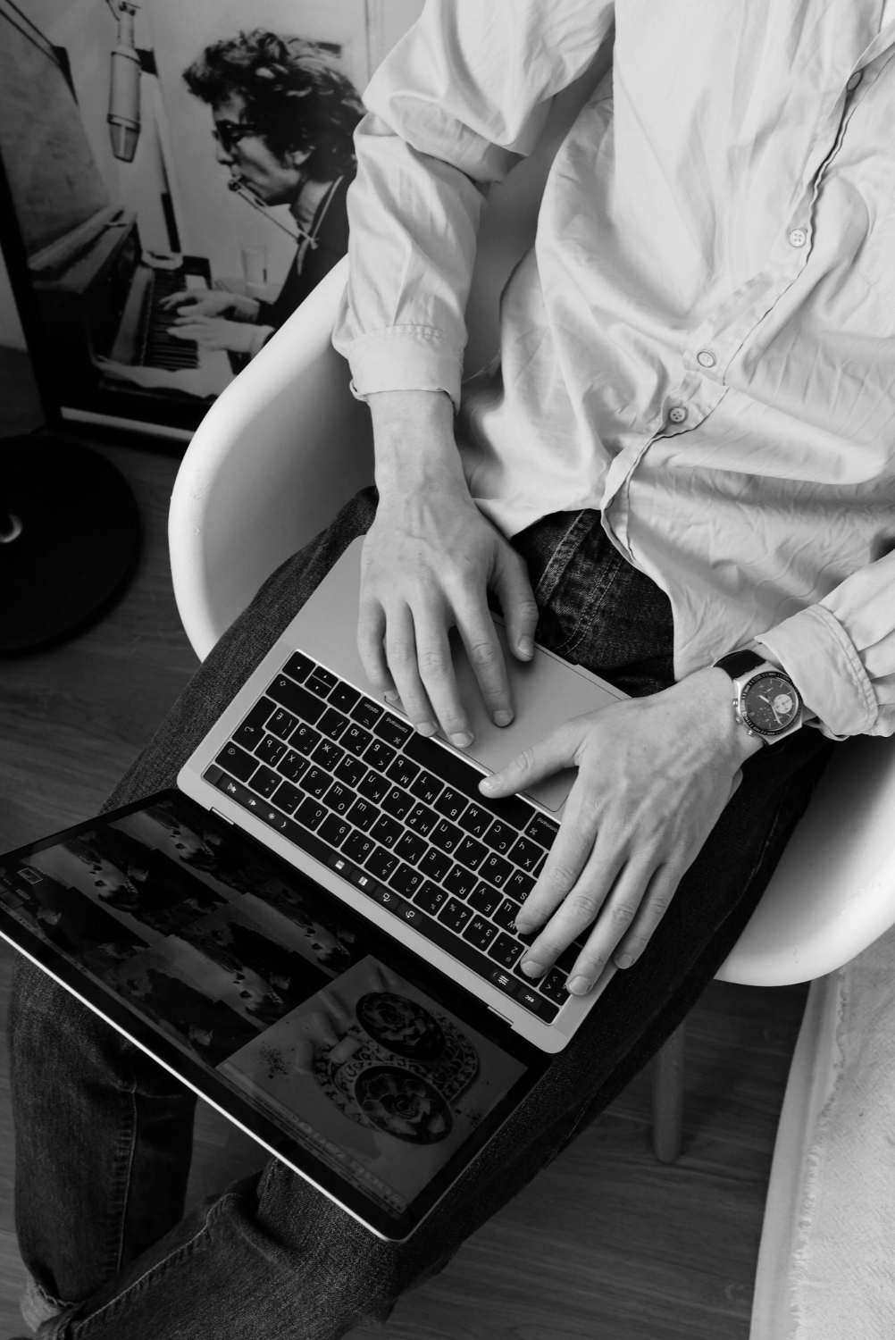 A black-and-white overhead photo of a person sitting in a chair with a laptop on their lap, typing with both hands, wearing a casual button-down shirt and watch, with a poster of a musician visible in the background.