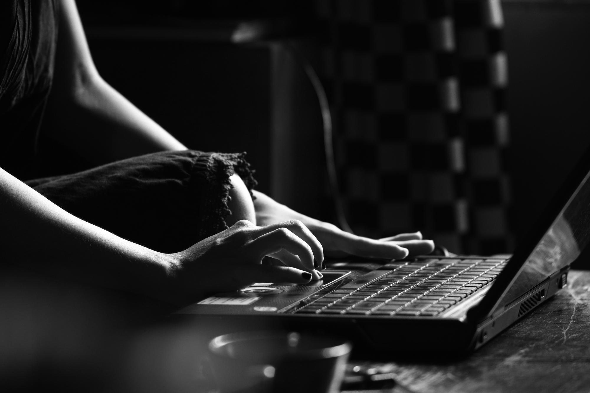 A black-and-white close-up of a person typing on a laptop, seated casually with one knee up, in a dimly lit room with a blurred background.